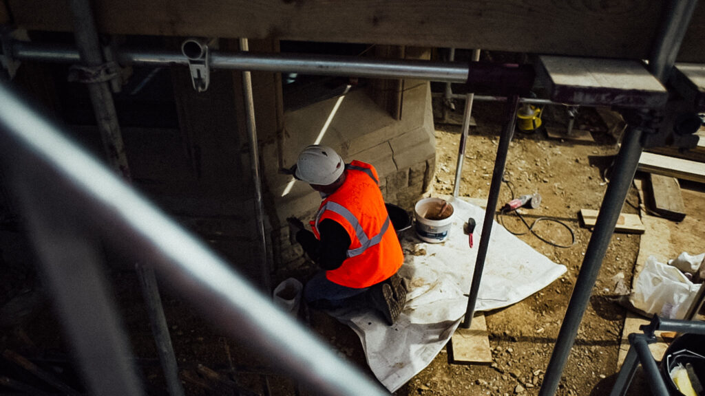 Downwards shot of a man working on a stone windowsill, surrounded by scaffolding. 