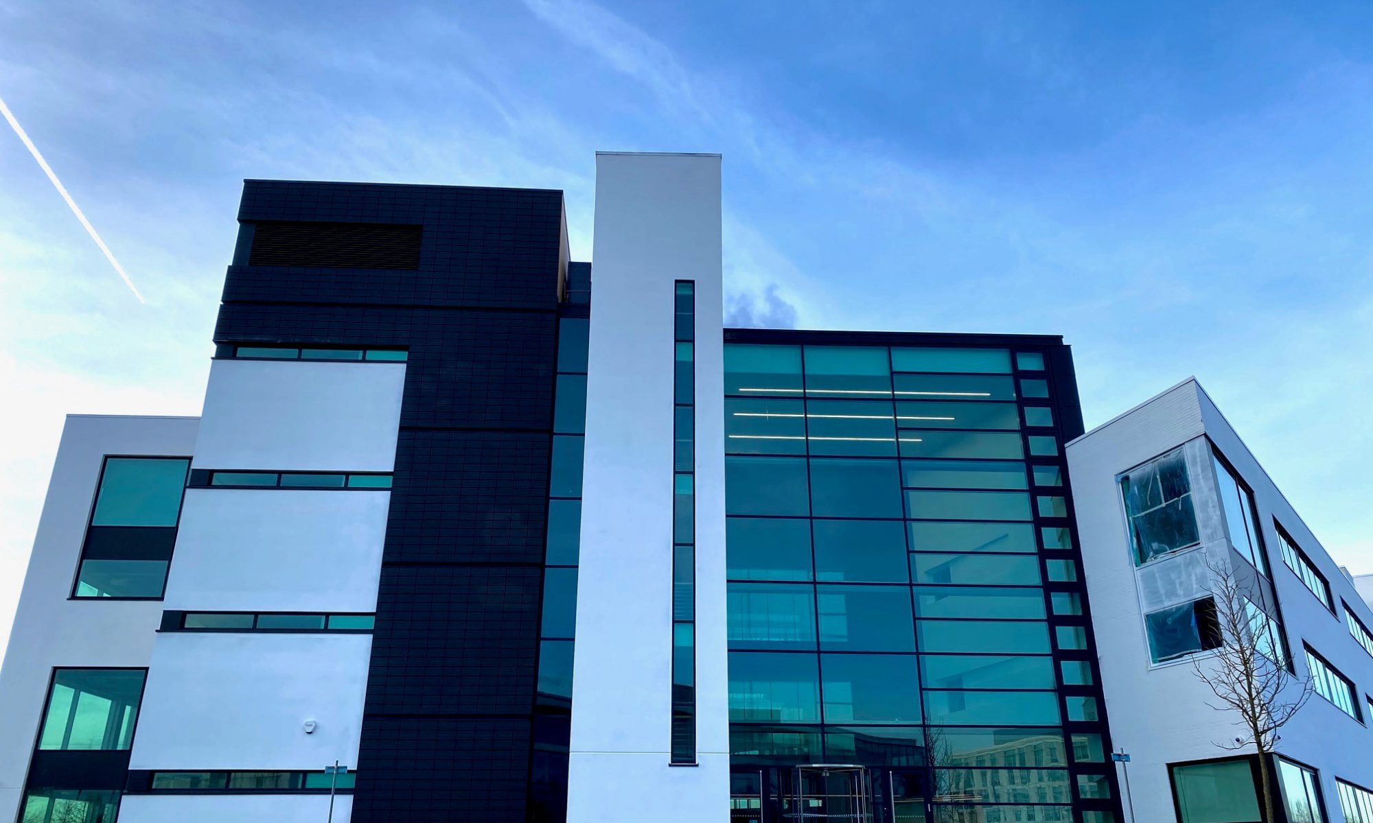 Modern glass office building with angular architecture, set against a vibrant blue sky with a streaking contrail.