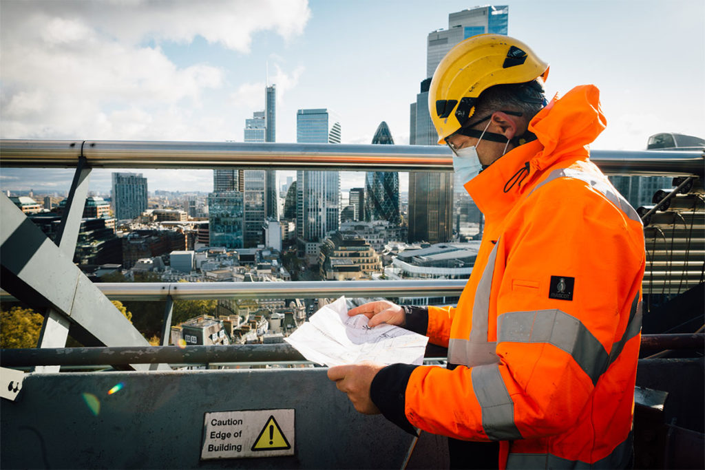 Construction worker in bright orange jacket and helmet examines city plans on rooftop, London skyline backdrop.