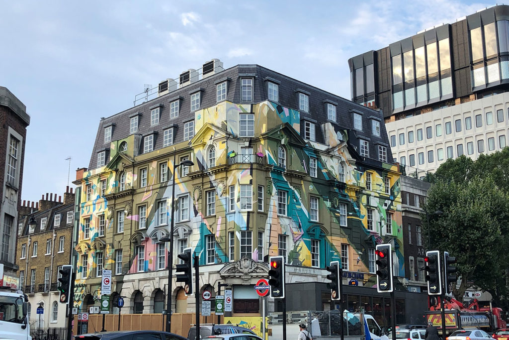 Vibrant geometric mural decorates large corner building in London, under cloudy sky at busy street junction.