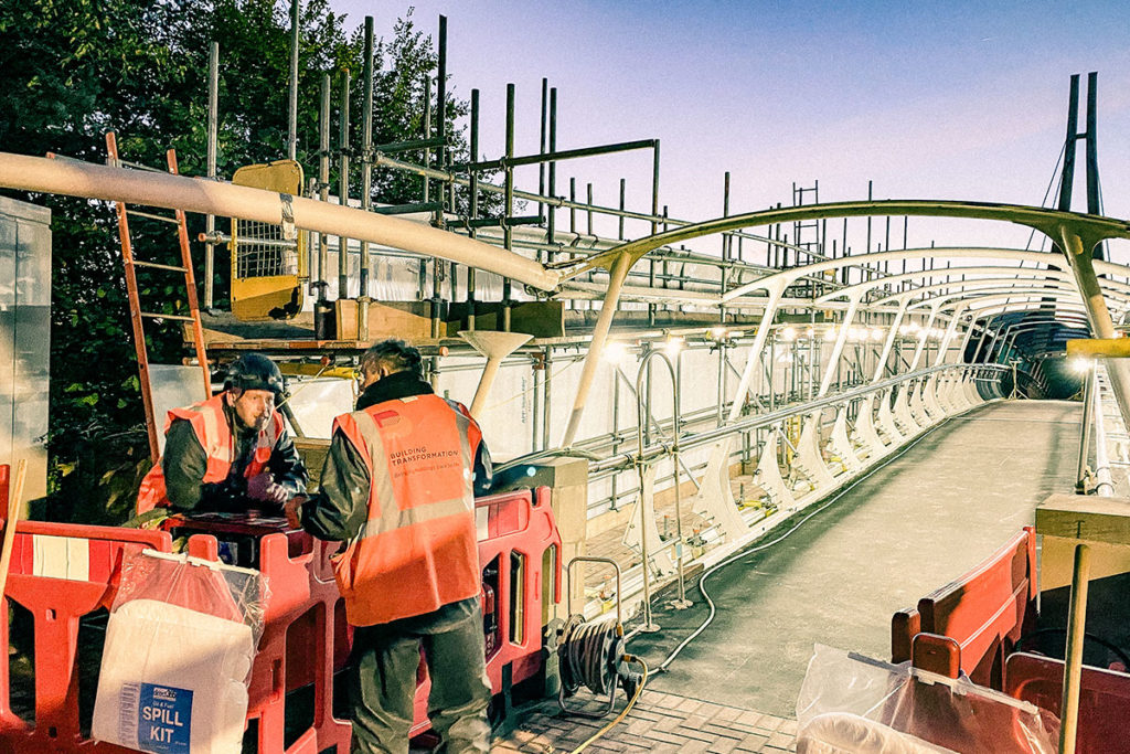 Construction workers in orange vests discuss progress beside a partially built bridge, lit under a dusky sky.