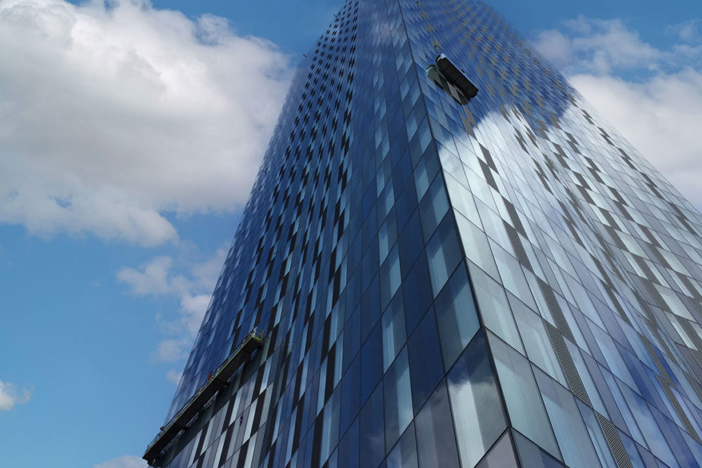 Modern glass skyscraper reflecting clouds, with suspended platforms for window cleaning against a blue sky.