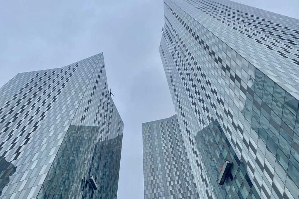 Tall modern glass skyscrapers with geometric patterns, viewed from below against a cloudy grey sky.
