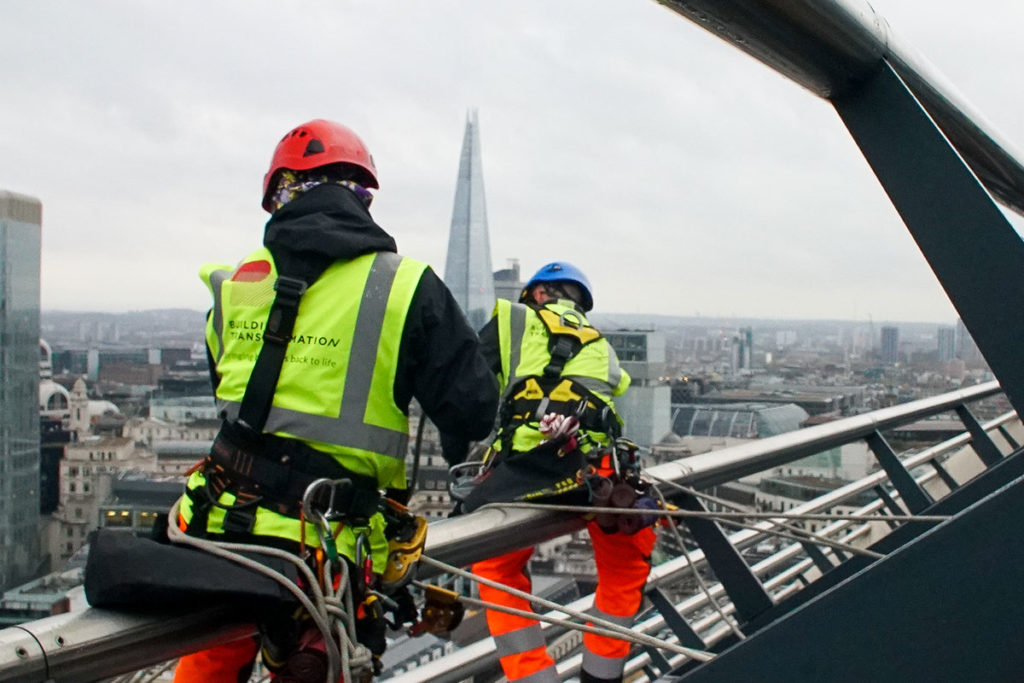 Construction workers in safety gear, atop a skyscraper, with London's skyline and the Shard in view.