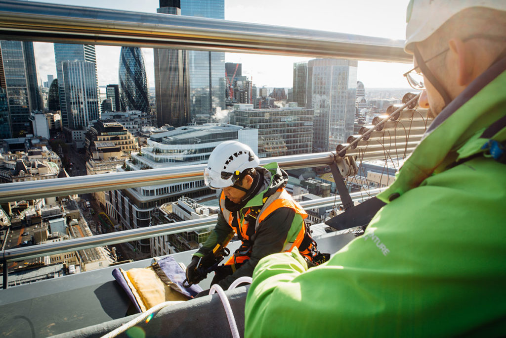 Engineers in protective gear inspect rooftop railings with London's skyline in the background.