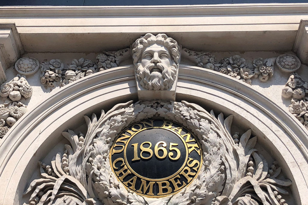 Ornate stone façade with Union Bank Chambers 1865 in gold, crowned by a sculpted bearded face.