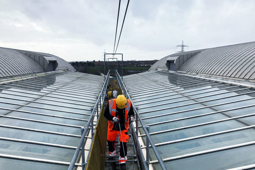Construction worker in orange vest and hard hat walks on scaffolding atop large industrial building.