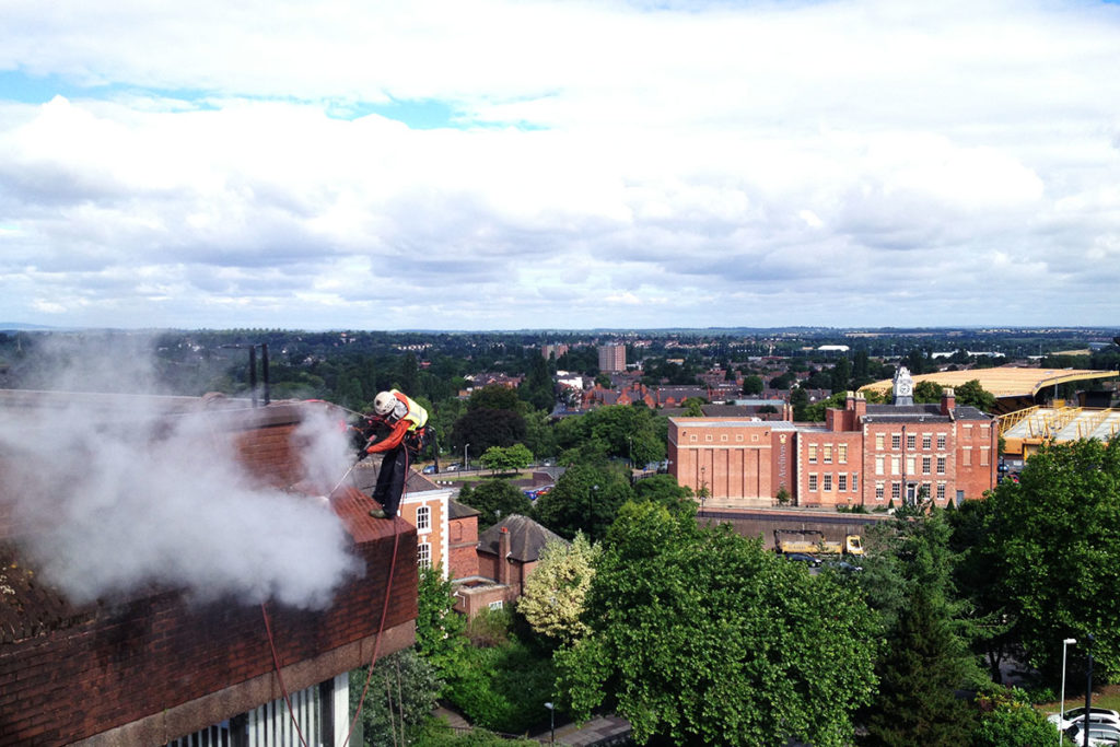 Rope access technician cleans smokey chimney on a rooftop, overlooking a scenic cityscape.