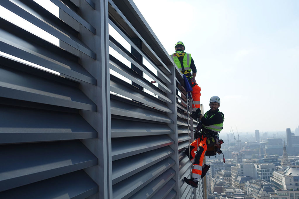 Rope access technicians suspended on a high-rise facade, wearing safety gear, cityscape in the background.
