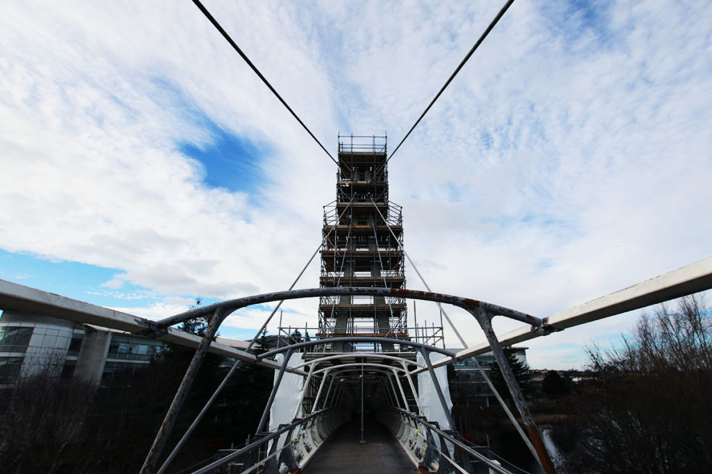 Scaffolding on a modern suspension bridge under a partly cloudy sky, with urban buildings in the background.