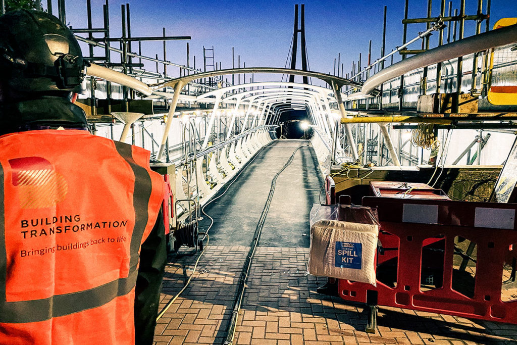 Construction worker in high-vis vest oversees illuminated bridge revitalisation project, with Spill Kit visible.