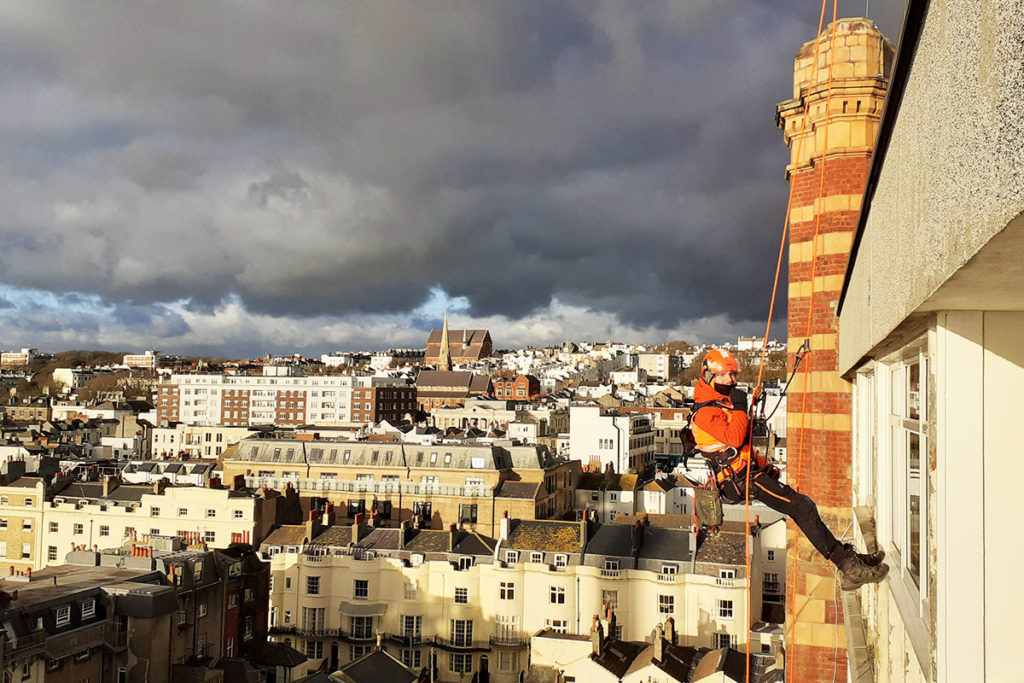 Rope access worker in orange gear abseils down building, cloudy sky and urban landscape in background.