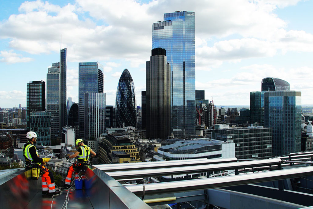 Workers in safety gear on a rooftop in front of London's iconic skyscrapers under a bright, cloudy sky.