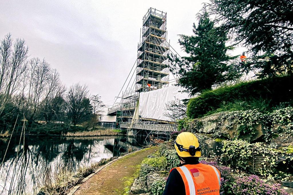 Construction site by a pond with scaffolding and worker wearing Building Transformation vest.