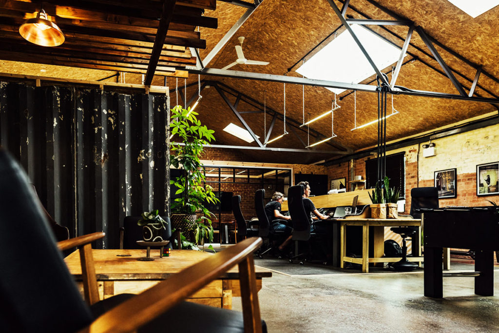 Spacious industrial-style co-working space with natural light, wooden beams, green plants, and focused workers.
