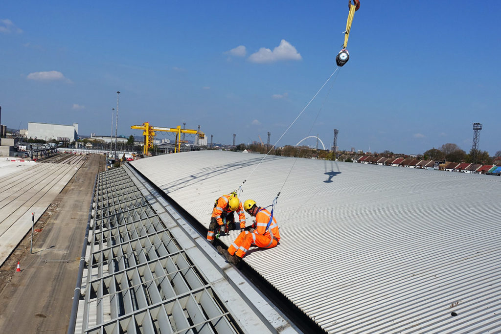 Two workers in bright orange safety gear inspecting a large industrial metal roof under a clear blue sky.