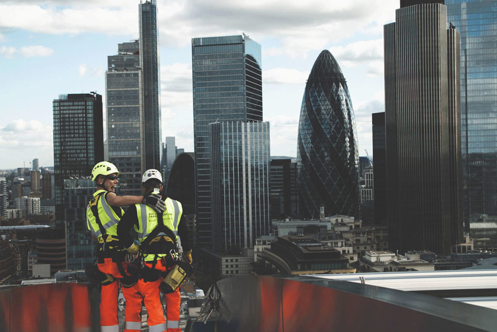 Construction workers in safety gear on rooftop overlooking London's skyline, featuring the Gherkin.
