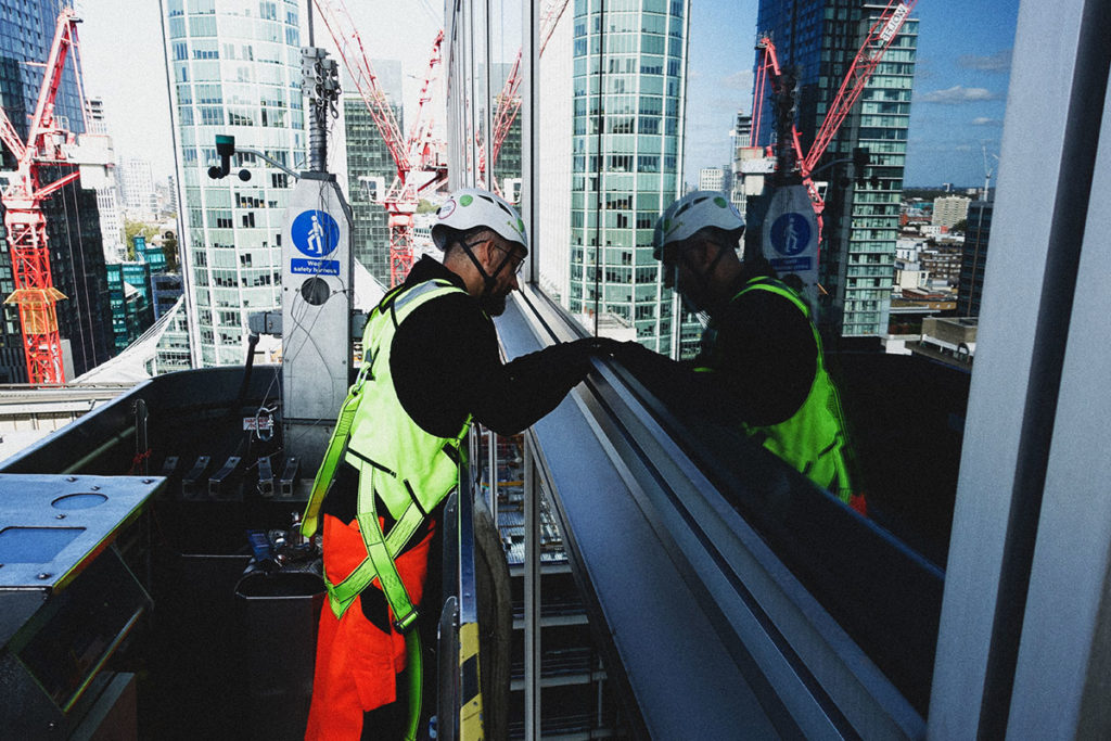 Construction worker in high-visibility gear inspects a skyscraper window with cityscape and cranes behind.