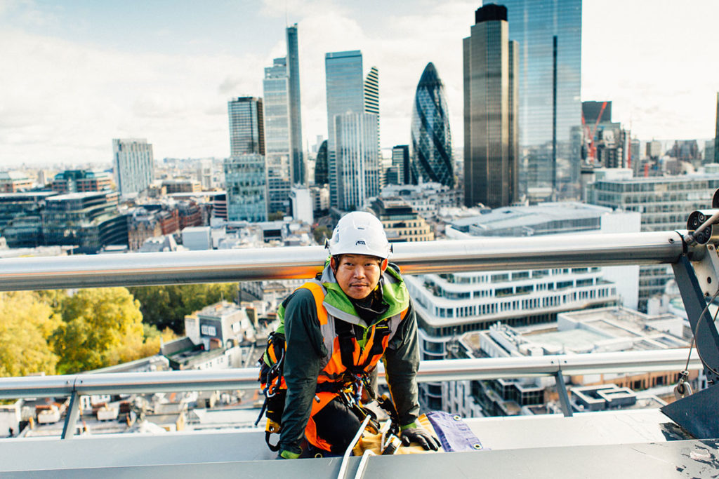 Construction worker in high-vis gear on rooftop with London skyline backdrop, including the Gherkin.