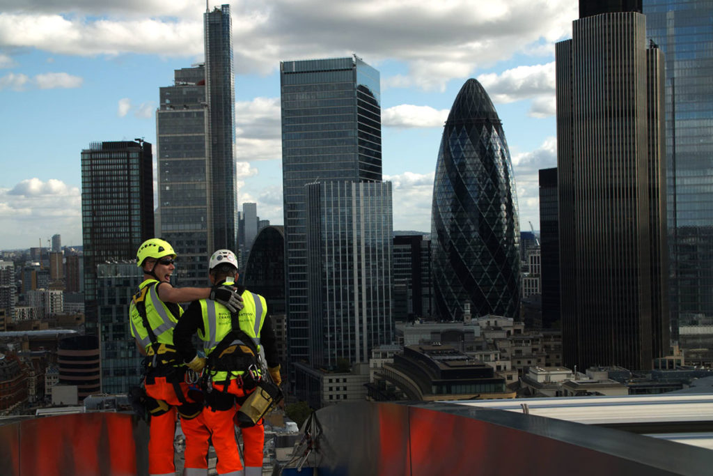 Construction workers in bright gear on London rooftop with city skyline and Gherkin building in view.