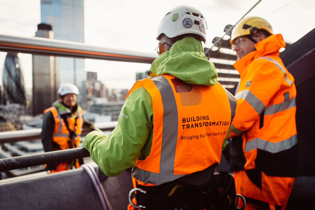 Construction workers on a high-rise, wearing orange vests with Building Transformation text, cityscape view.