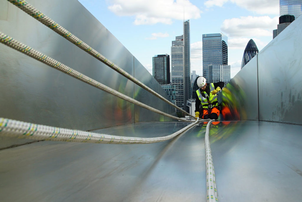 Construction worker in safety gear securing ropes on a rooftop with London skyline in the background.
