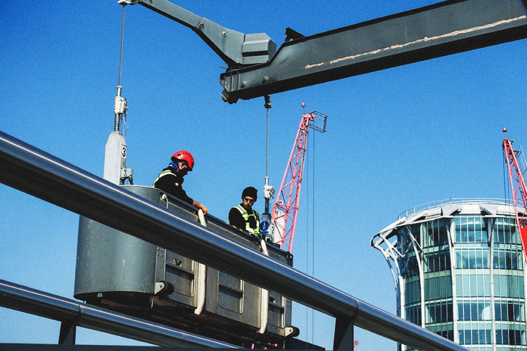 Two construction workers in harnesses on a platform, vivid blue sky, red cranes, modern glass building.