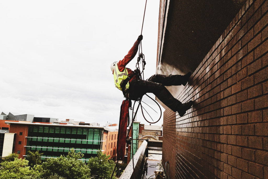 Worker in safety gear rappelling down a tall brick building, overlooking modern cityscape.