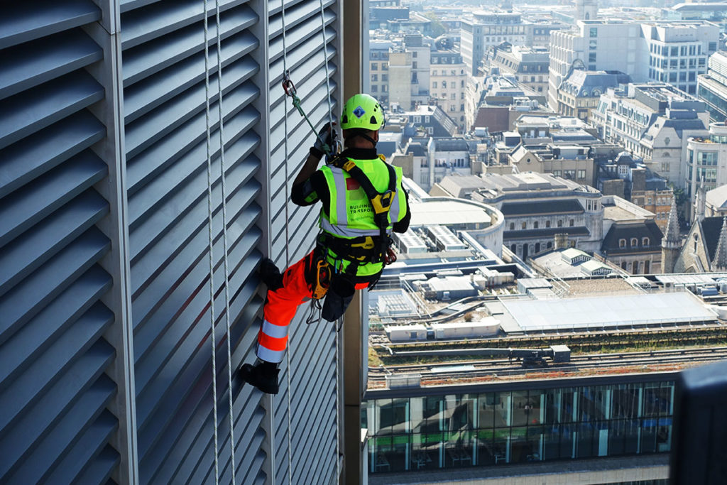 Safety worker with helmet and harness rappelling down building, urban skyline in background.