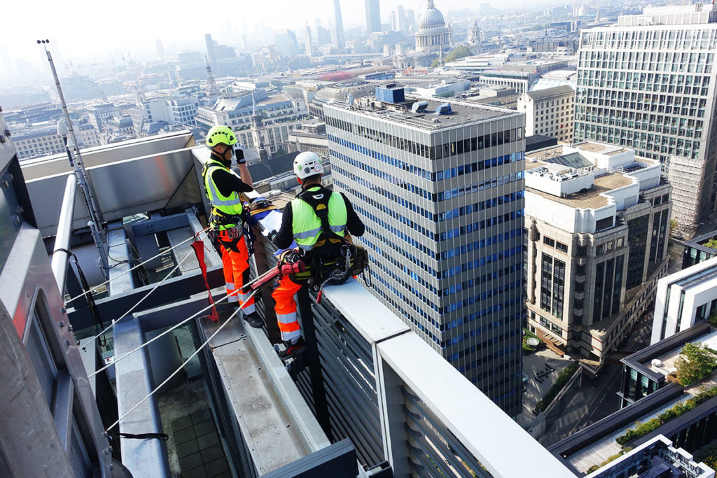 Two workers in safety gear perform maintenance on a skyscraper rooftop, overlooking a cityscape.