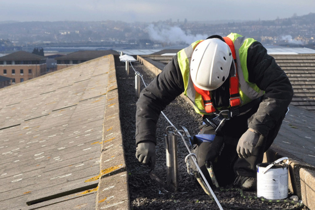 Worker in safety gear installing rooftop safety cables; urban skyline visible in the background.