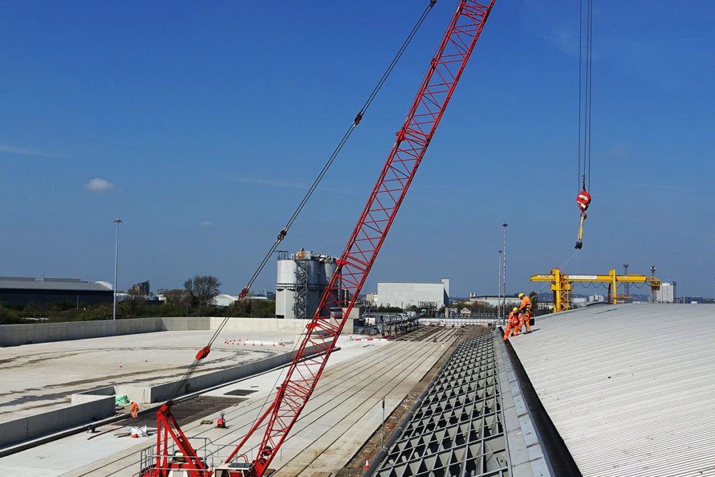 Large red crane over railway construction with workers in orange gear under a clear blue sky.