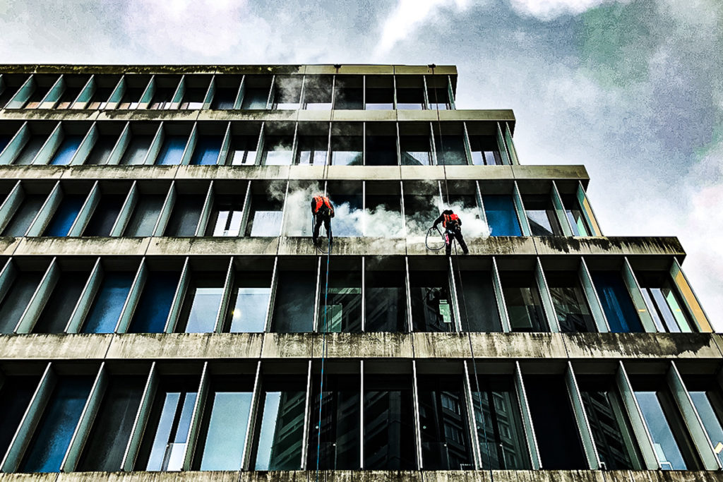 Window cleaners in red vests abseil down a glass building, surrounded by clouds of detergent.