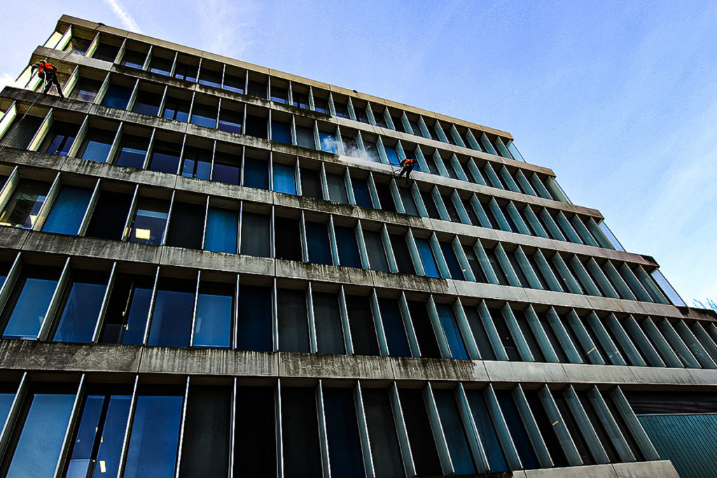 Window cleaners in safety gear abseiling down a modern glass building against a clear blue sky.