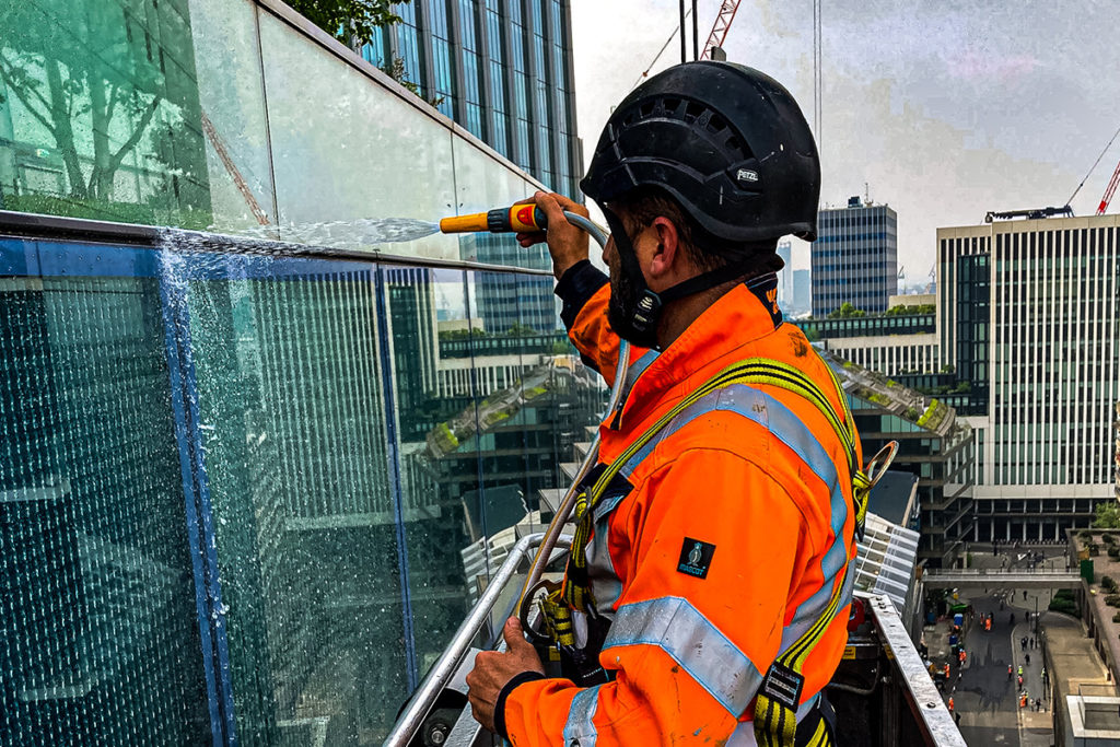 Construction worker in orange safety gear cleaning skyscraper windows with a hose on a raised platform.