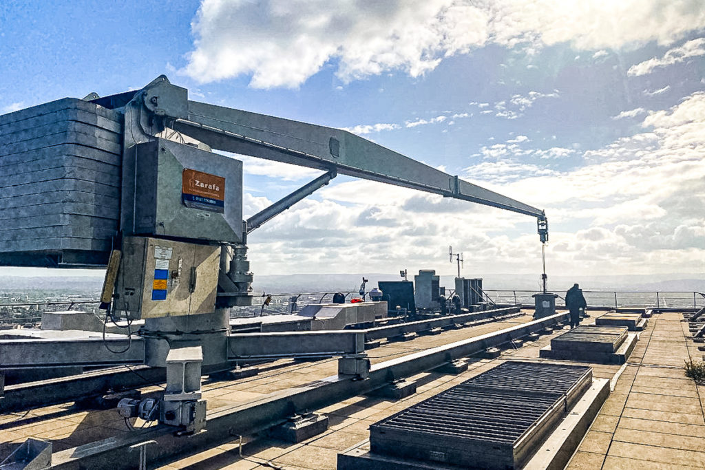 Industrial crane labelled Zarafa on a sunny rooftop, silhouetted worker, and blue sky with clouds.