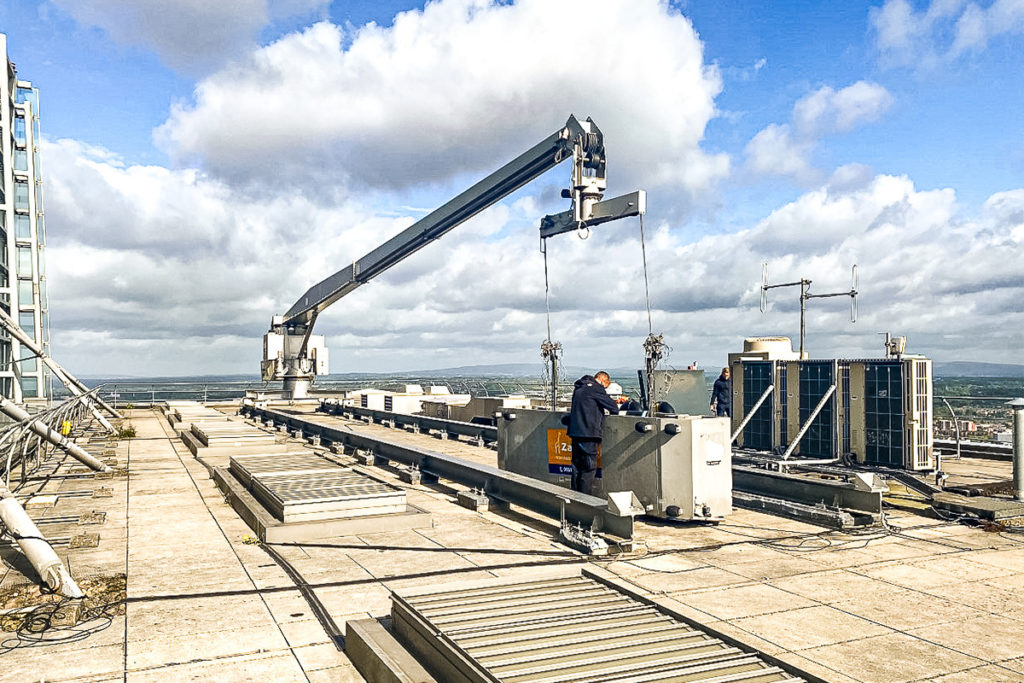Workers inspect roof-mounted crane on building under cloudy sky, with solar panels in background.