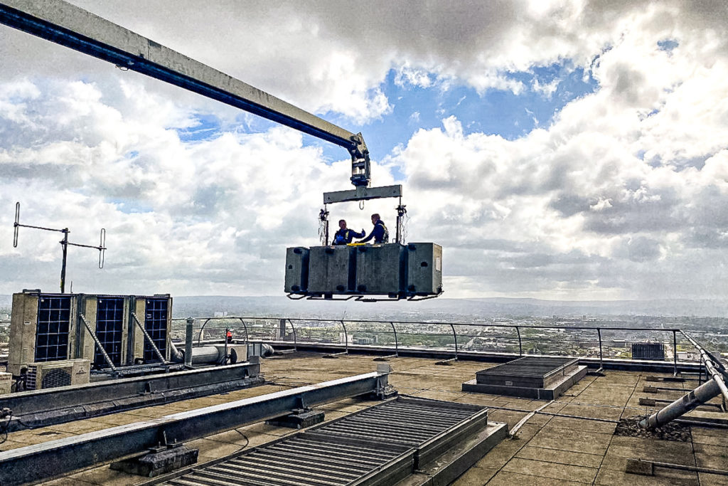Workers securing platform on building rooftop under cloudy sky, urban landscape in background.