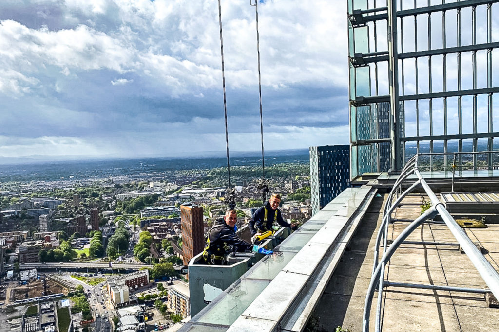 Window cleaners suspended on a skyscraper rooftop, with a sprawling cityscape under a cloudy sky.