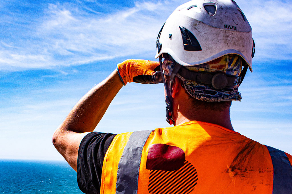 Construction worker in safety gear surveying the ocean, wearing an orange vest and white helmet in sunlight.