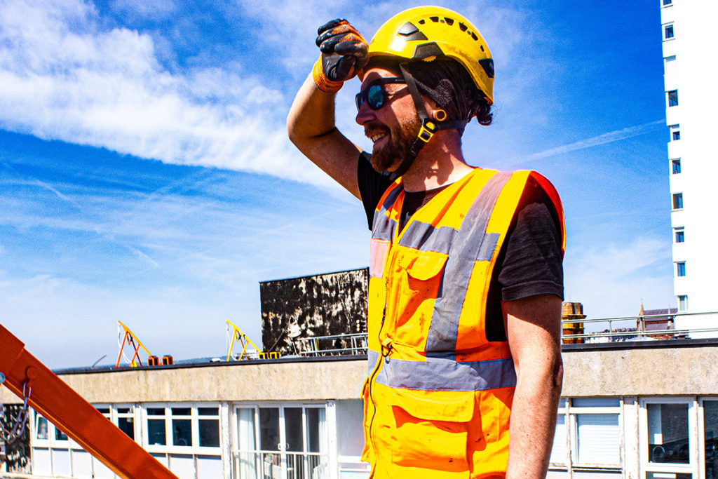 Construction worker in hi-vis vest and helmet, squinting under bright sun on a building rooftop.