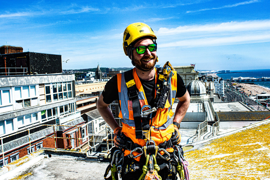 Construction worker in safety gear on a rooftop with a seaside cityscape in the background, sunny day.