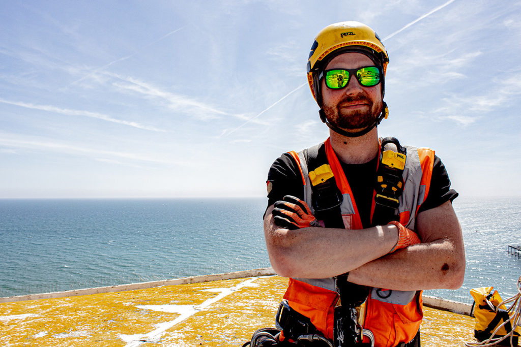 Worker in bright safety gear and helmet stands confidently by the sea under a clear blue sky.