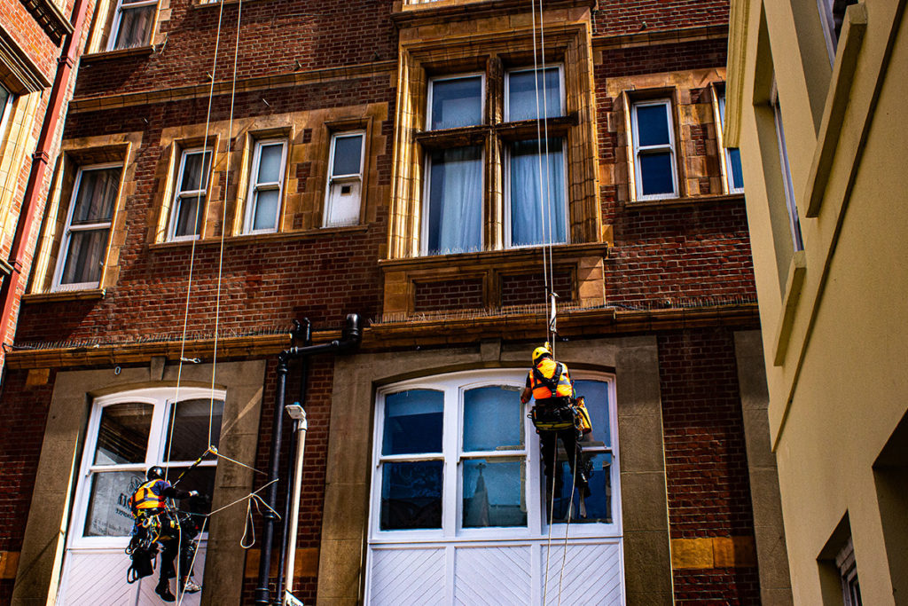 Two workers in safety gear suspended on ropes clean windows of a red brick building under bright daylight.