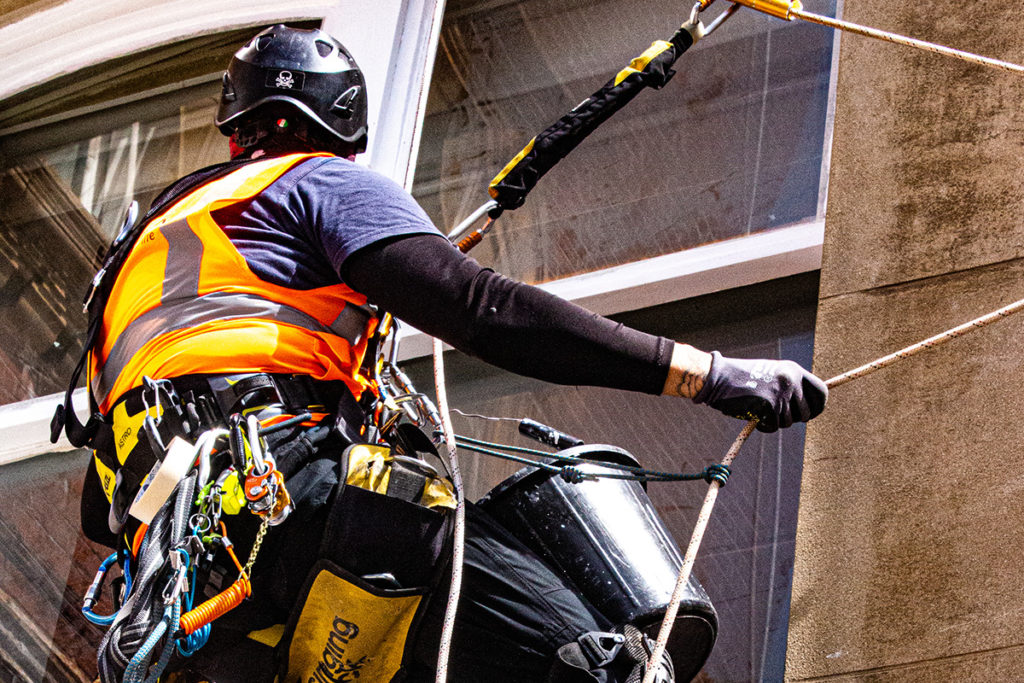 Window cleaner in safety gear, wearing a harness and helmet, operates on a high-rise building wall.