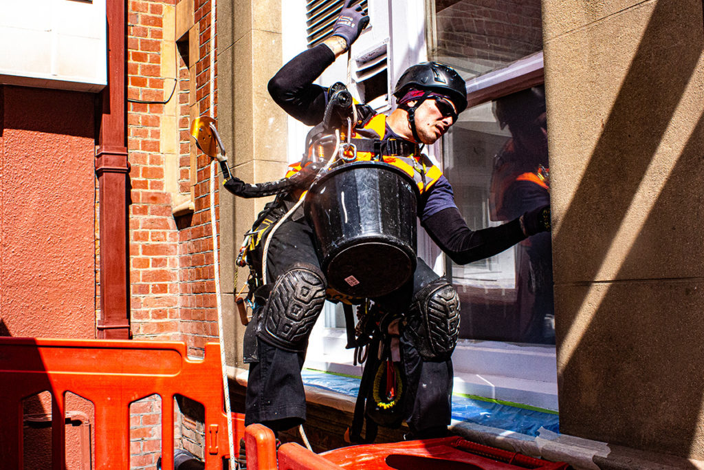 Worker in climbing gear cleans building window with bucket and gear, sunlight casts strong shadows.