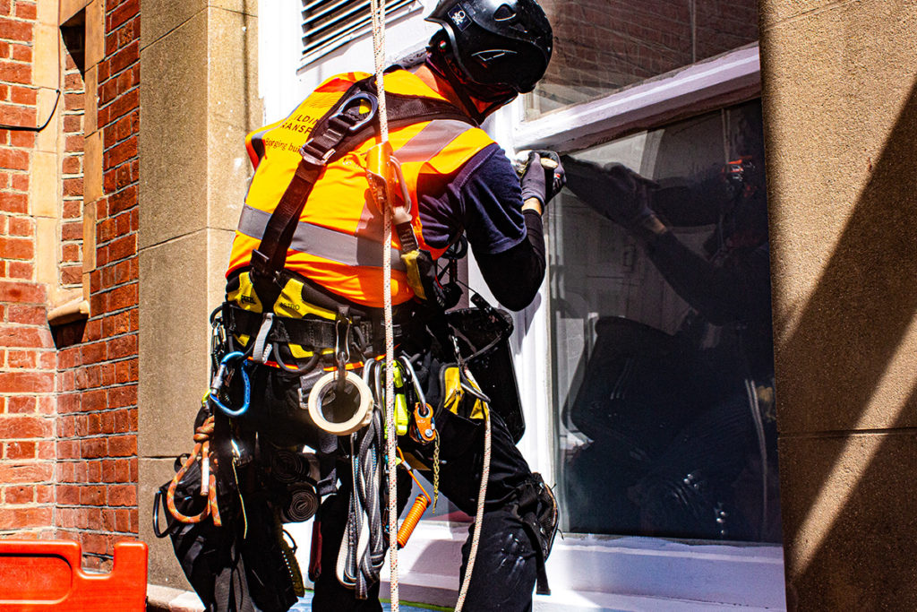 Construction worker in high-vis gear repairing windows on a brick building with safety harness and tools.