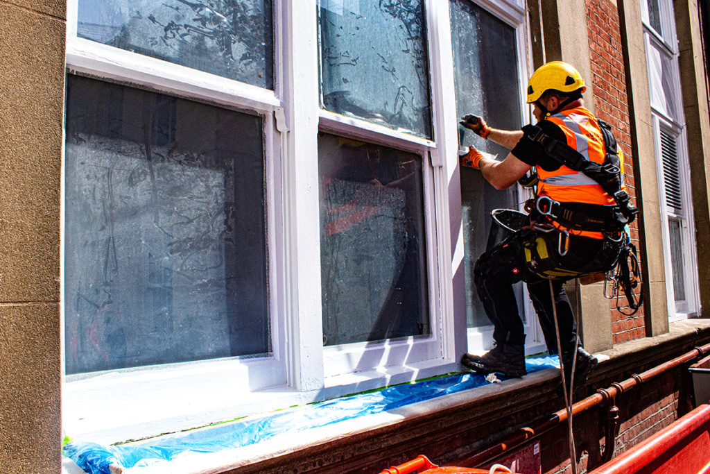 Window cleaner in safety gear scales building facade, cleaning large windows on sunny day.