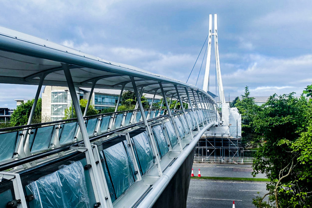 Modern white pedestrian bridge with tall pylons, glass panels, and lush greenery under a cloudy sky.