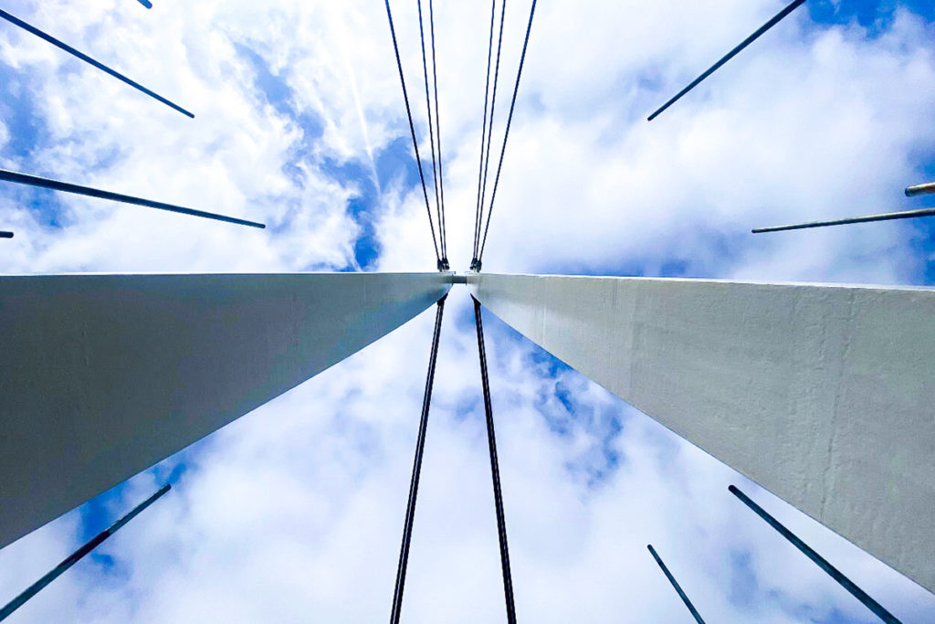 Modern suspension bridge cables against a bright, cloudy sky, viewed from below, conveying grandeur.